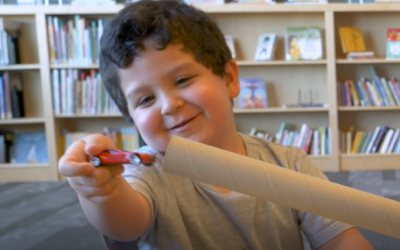 young boy in library playing with toy car, about to put car inside long cardboard tube